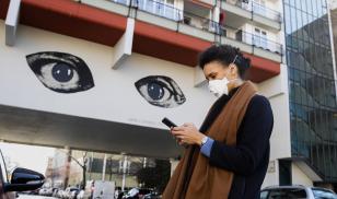 A young woman wearing a protective mask looks at her smartphone while passing by a grafitti representing two big watching eyes in Berlin, Germany on April 1, 2020. Illustrative Editorial (Photo by Emmanuele Contini/NurPhoto via Getty Images)